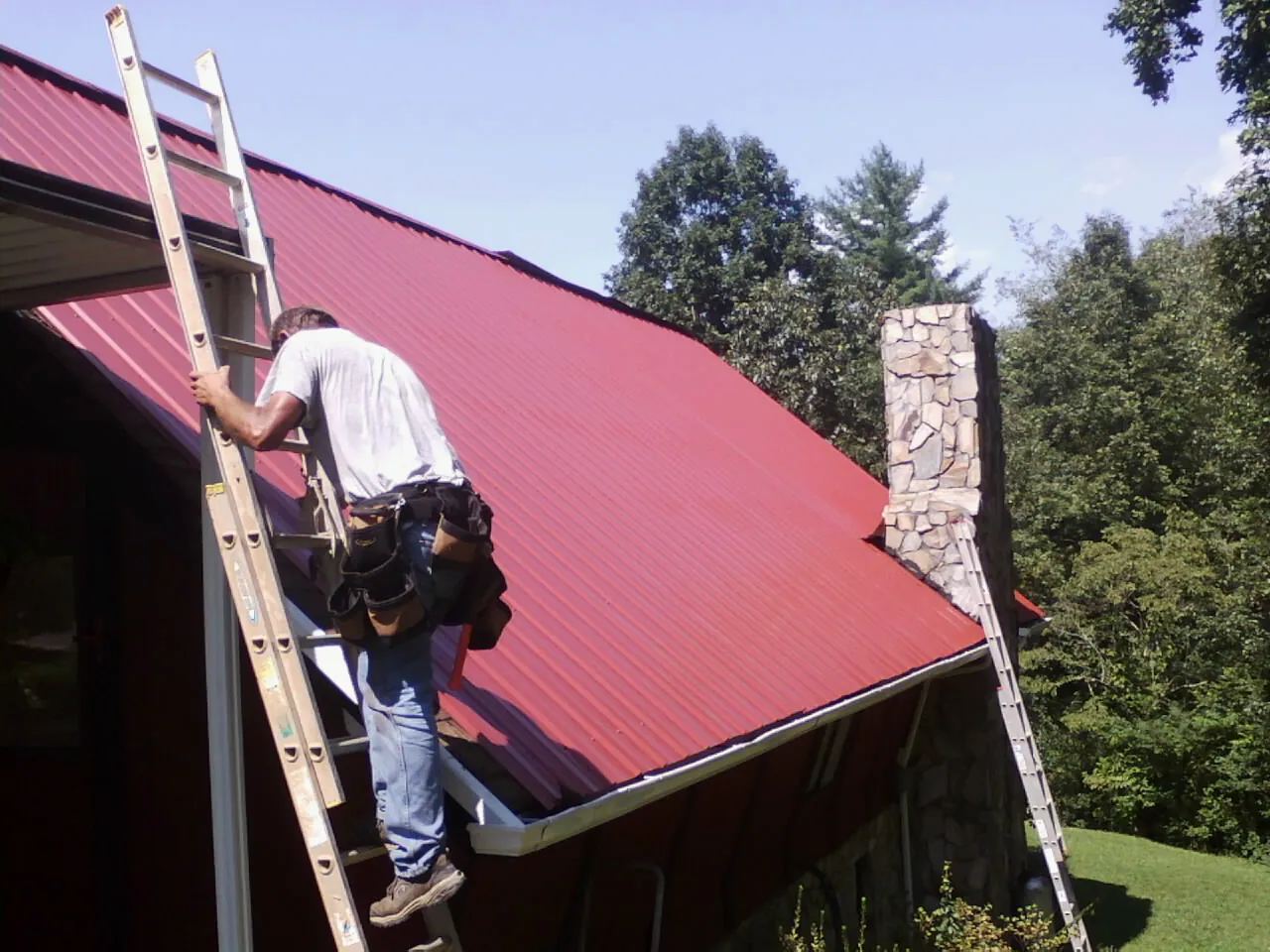 Roofer on a ladder beside a red metal roof and stone chimney — Soffit & Fascia Repair by Roofing Brothers in Menahga